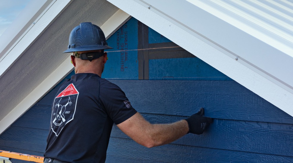 Un travailleur portant un casque, un t-shirt et des gants applique des moulures extérieures sous le sommet d’une ligne de toit.