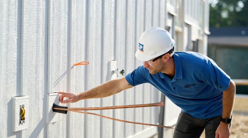 A worker in a hard hat and t shirt inspecting the exterior side of a siding clad building where some wires are protruding.