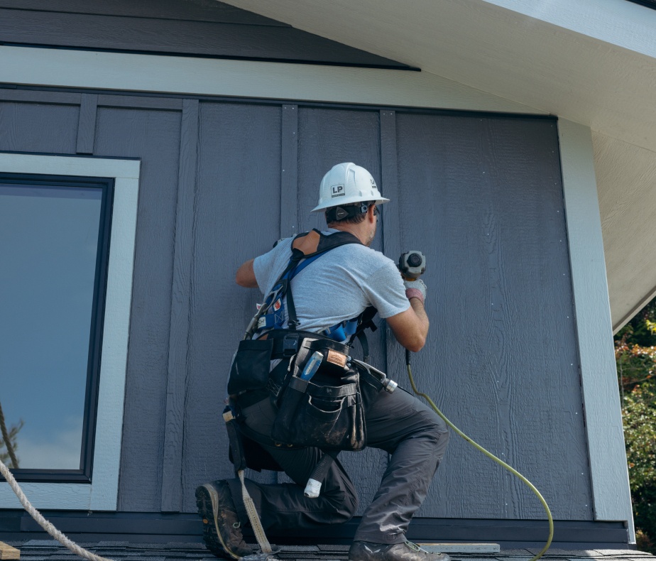 A worker in a hard hat up a ladder applying vertical siding to the side of a house.