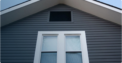 Mirando el costado de una casa hacia la línea puntiaguda del techo, con un revestimiento horizontal oscuro y una ventana con molduras blancas debajo.